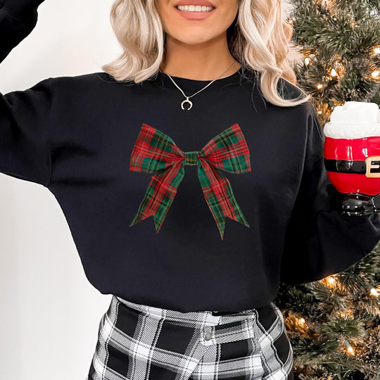Model wearing a black crewneck sweatshirt with a red and green plaid bow graphic, holding a Santa mug