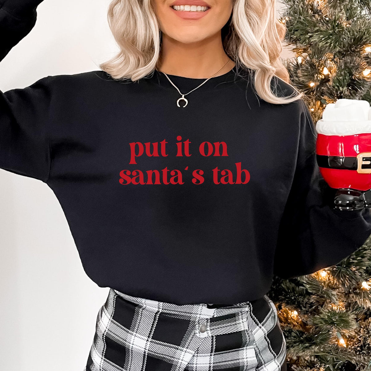 Model wearing a black crewneck sweatshirt with put it on santa's tab in red lettering, holding a Santa mug