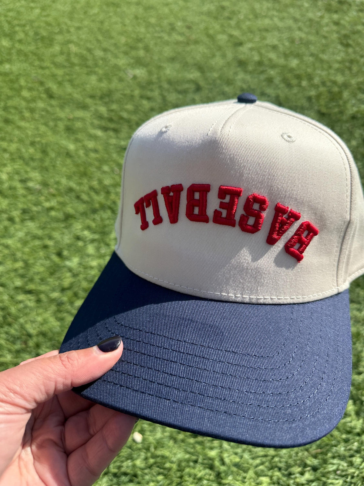 Hand holding the upside-down Baseball snapback hat, natural front with navy brim and red arched lettering, on grass