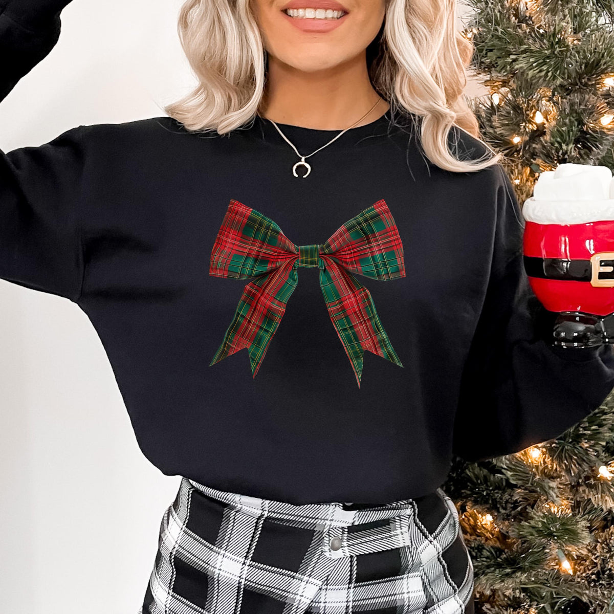 Model wearing a black crewneck sweatshirt with a red and green plaid bow graphic, holding a Santa mug