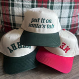 Put It on Santa's Tab snapback in green with two upside-down Merry hats against a plaid pillow