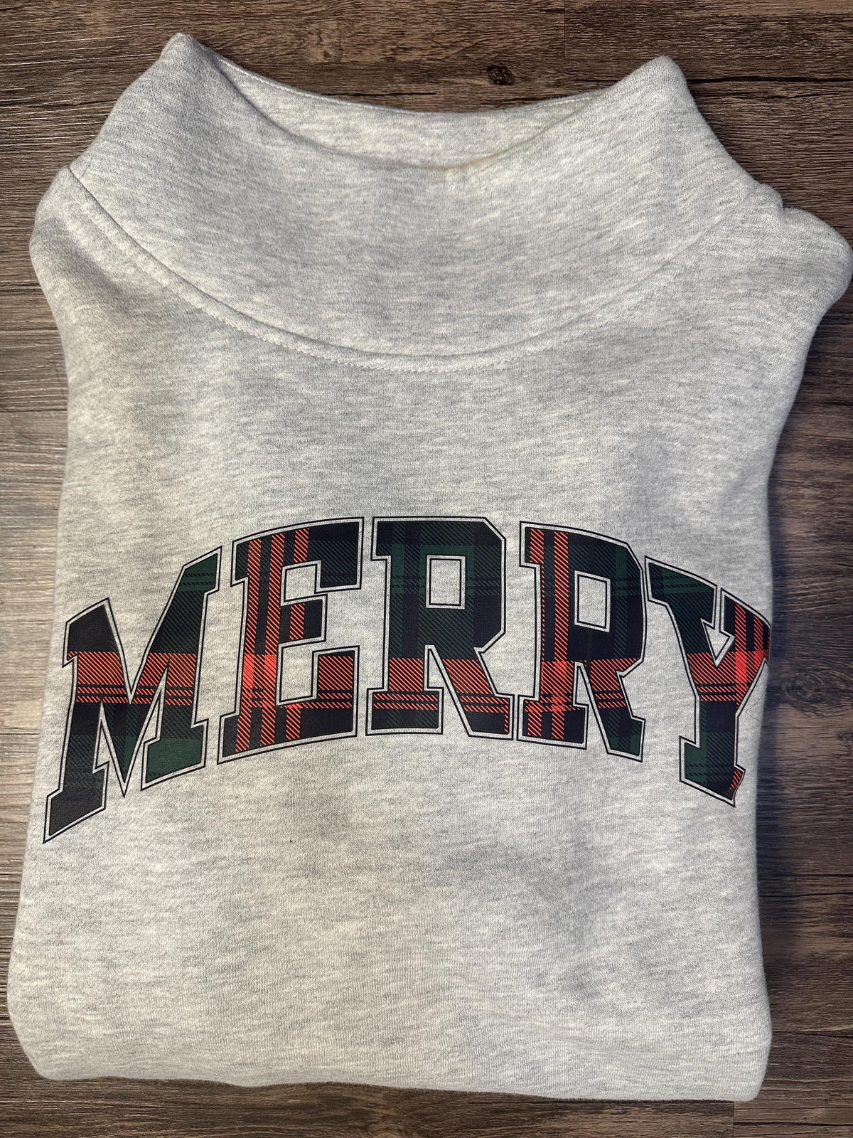 Grey mockneck sweatshirt with MERRY in red and green plaid varsity letters, folded flat