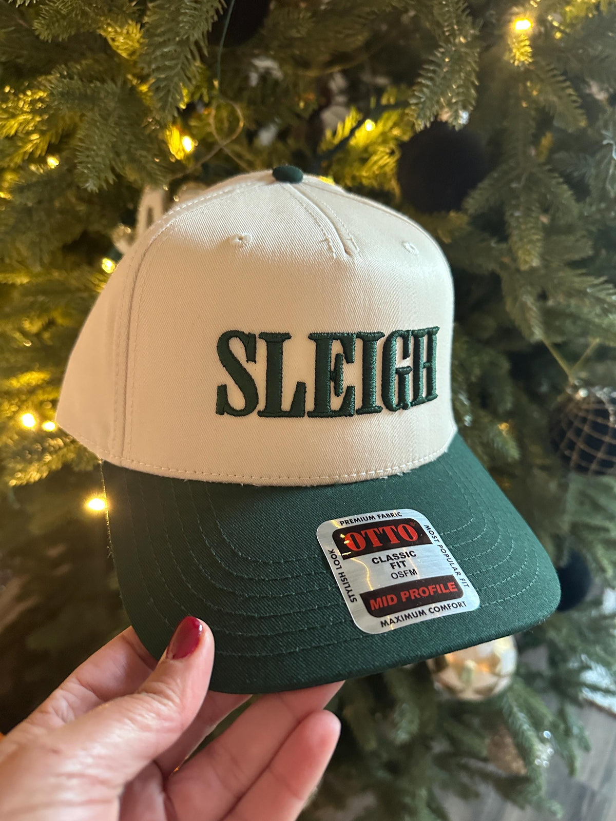 Hand holding the Sleigh snapback hat in natural with a green brim and green lettering by a Christmas tree